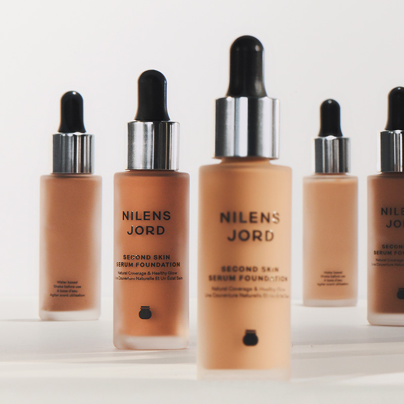 Nilens Jord Second Skin Serum Foundation. Useita beige meikkivoidepulloja, joissa on musta pipetti ja hopeanvärinen korkki. Pulloissa on mustaa tekstiä.