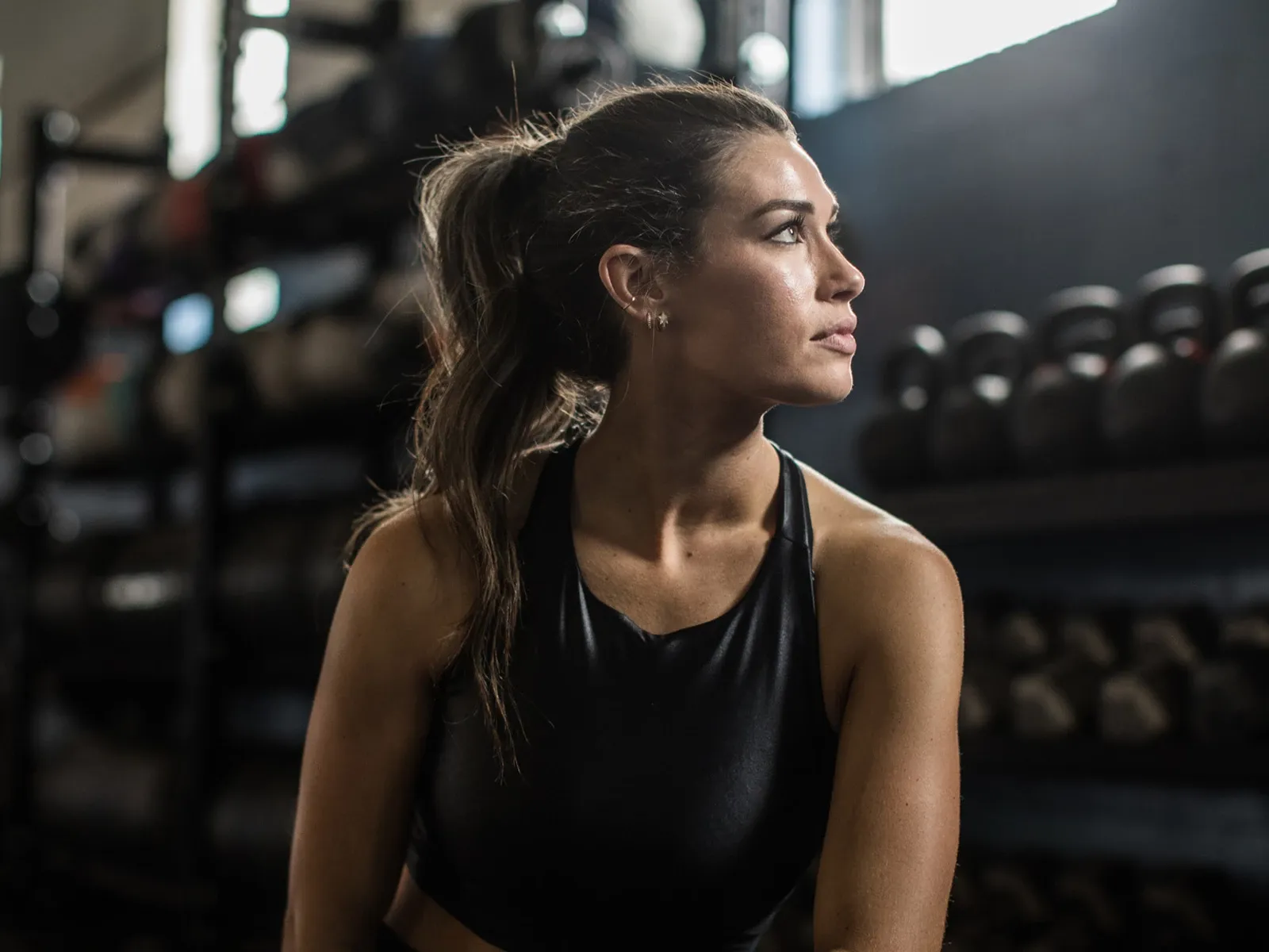 Kvinne i treningsklær sitter på treningssenter og ser til siden, med kettlebells i bakgrunnen.