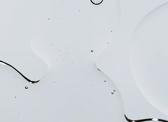 Nærbilde av en gjennomsiktig hudpleietekstur som flyter ut i myke former mot lys bakgrunn, med små dråper og tynne kanter av serum.