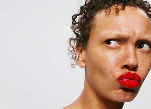 Person med röda läppar och naturlig makeup som tittar åt sidan med rynkad panna, med små fräknar och ett litet hjärta ritat på kinden mot ljus bakgrund.