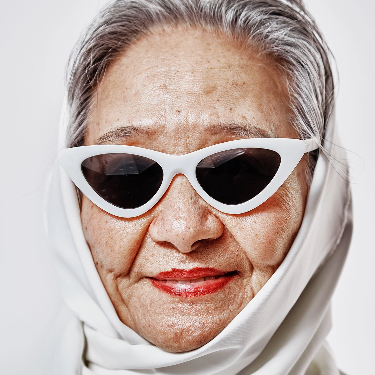 Närbild av en person som bär vita solglasögon och rött läppstift, med fokus på hud och läppar i en minimalistisk, ljus bakgrund.