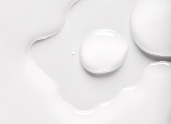 Lähikuva valkoisista nesteen pisaroista vaalealla pinnalla, pehmeitä muotoja ja heijastuksia.