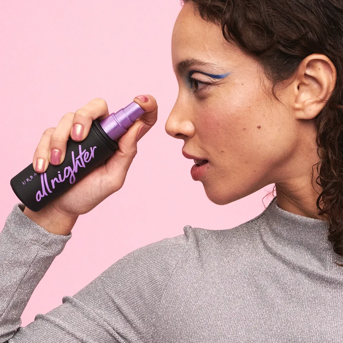 En kvinna med lockigt hår och makeup i profil, som visar upp en All Nighter Setting Spray från Urban Decay, mot en rosa bakgrund.