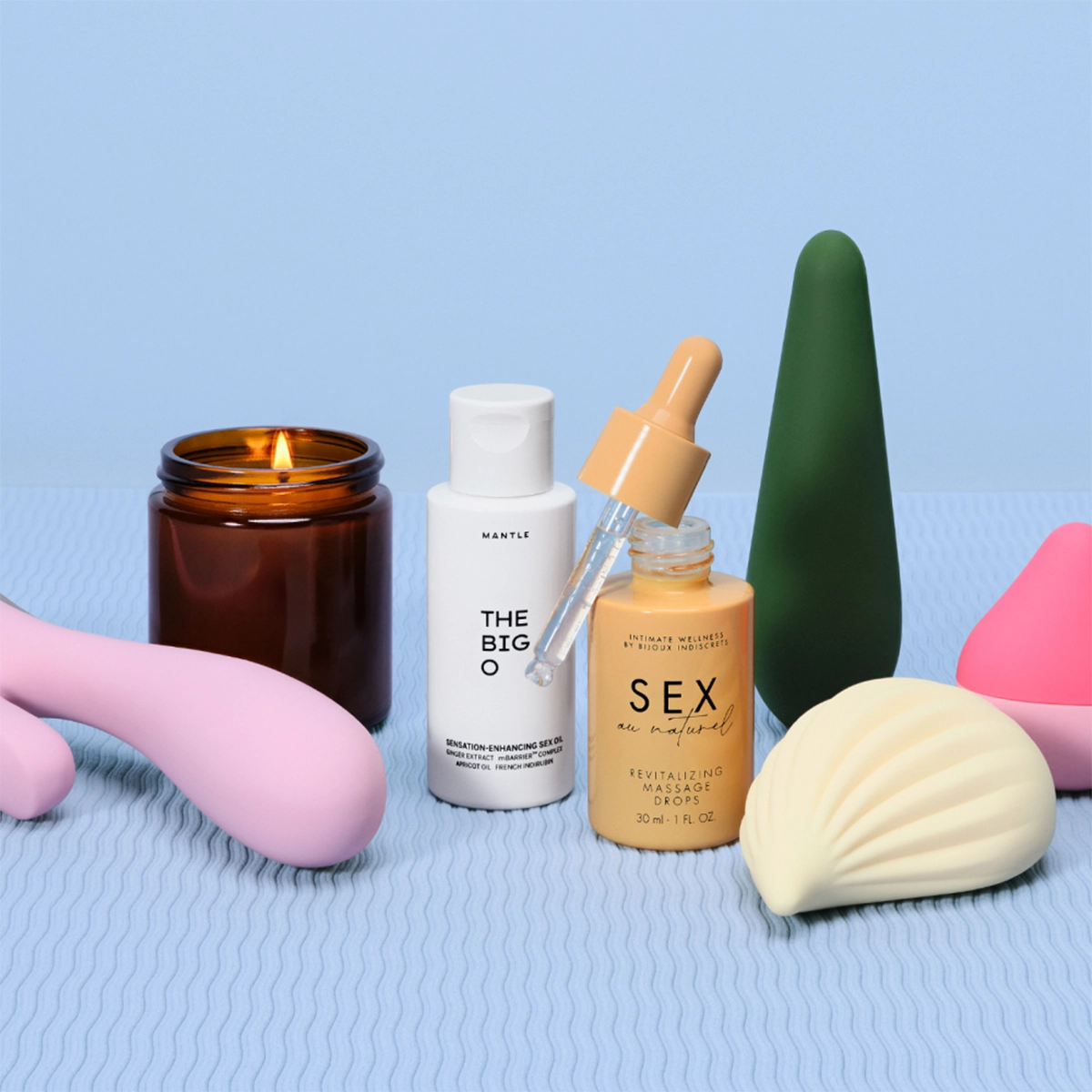 Asetelma intiimituotteista ja vartalonhoidosta vaaleansinisellä taustalla. Palava tuoksukynttilä on sijoitettu Mantle The Big S*xcation Enhancing Serum -seerumin ja “Sex Oil” revitalizing massage oil -hierontaöljyn viereen, mukana pinkkejä ja vihreitä seksileluja.