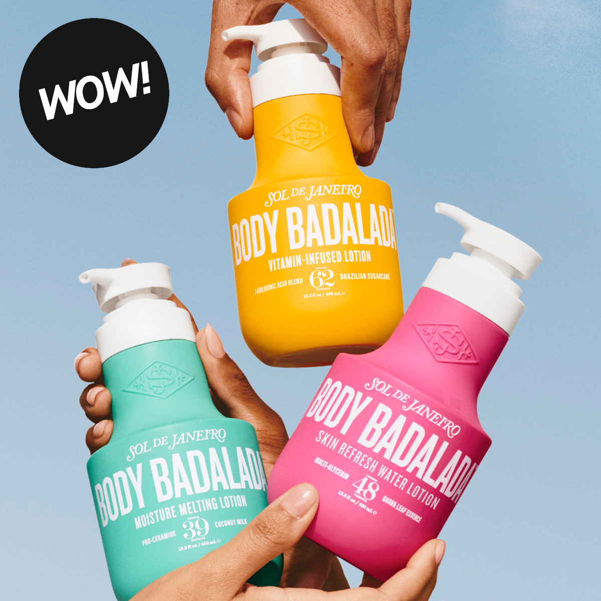 Tre Sol de Janeiro Body Badalada body lotions hålls upp av händer mot en blå himmel. Förpackningarna visar texterna "Body Badalada", "Moisture Melting Lotion Cheirosa 39 Coconut Milk", "Vitamin-Infused Lotion Cheirosa 62 Brazilian Sugarcane" och "Skin Refresh Water Lotion Cheirosa 48 Guava Leaf Essence", alla 400 ml, samt en svart etikett med texten "WOW!" i övre vänstra hörnet.