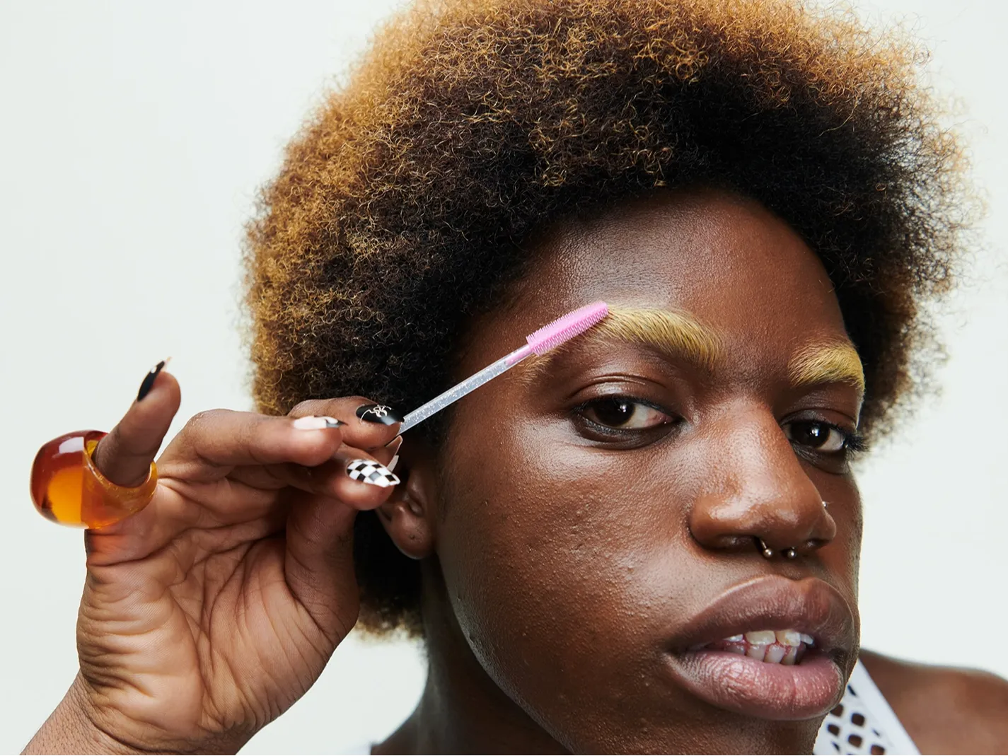 Närbild av en person som applicerar bryngel med en rosa spoolieborste på blekta ögonbryn, med fokus på ögonbrynsmakeup och hudens textur. Naturlig sminklook med markerade bryn och neutral bakgrund.