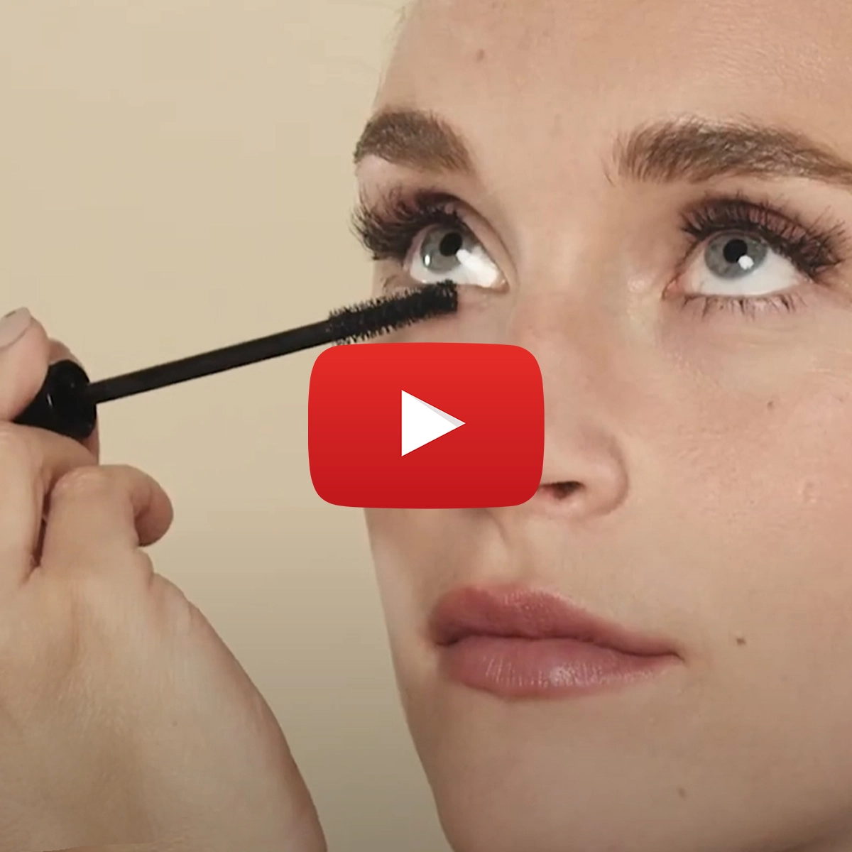 Person som tittar uppåt och applicerar mascara mot en beige bakgrund.
