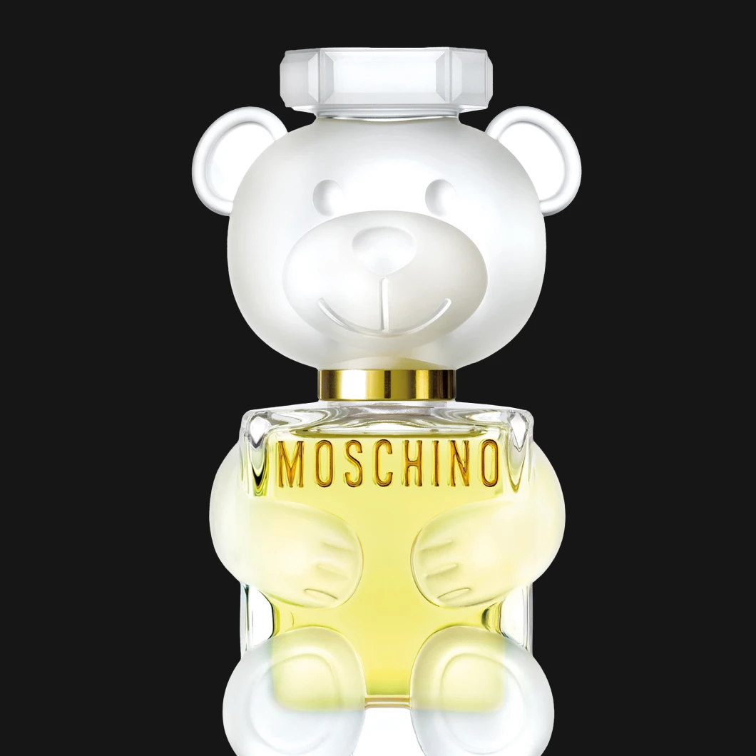 Moschino parfymeflaske formet som en teddybjørn i frostet glass, med lysegul væske, mot svart bakgrunn.