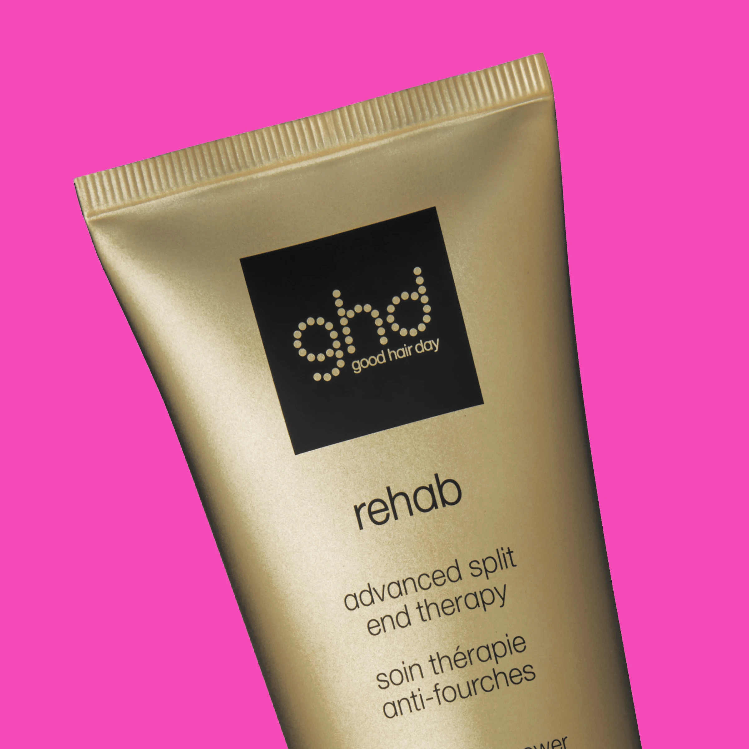 En gullfarget tube fra ghd med sort etikett mot rosa bakgrunn.