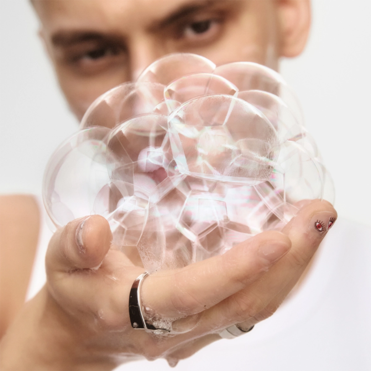 Närbild av en hand som håller en klunga genomskinliga såpbubblor framför ett ansikte i bakgrunden mot ljus bakgrund. Fokus ligger på de skimrande bubblorna och deras reflektioner.
