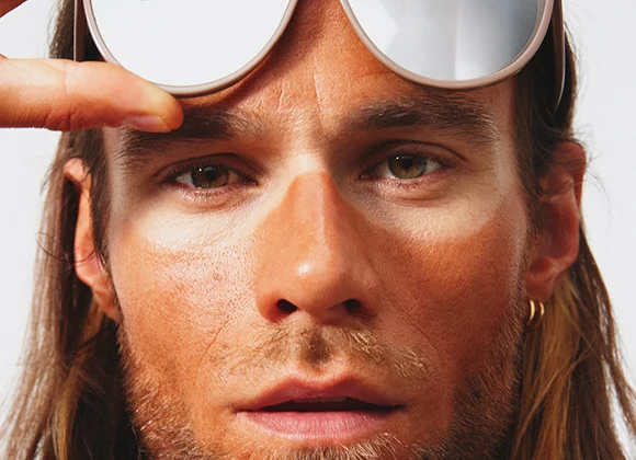 En person løfter et par store, speilblanke solbriller over øynene, noe som fremhever tydelige solbeskyttelseslinjer og kontrast mellom solbrune og beskyttede partier i ansiktet, med fokus på hudpleie og SPF.