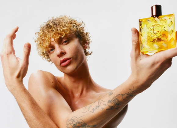 Henkilö, jolla on lyhyet kiharat hiukset, pitää kädessään keltaista hajuvesipulloa, jossa lukee "Versace Eros Energy" ja Versacen logo etupuolella. Läpinäkyvässä pullossa on kullanvärinen neste ja musta korkki vaaleaa taustaa vasten.