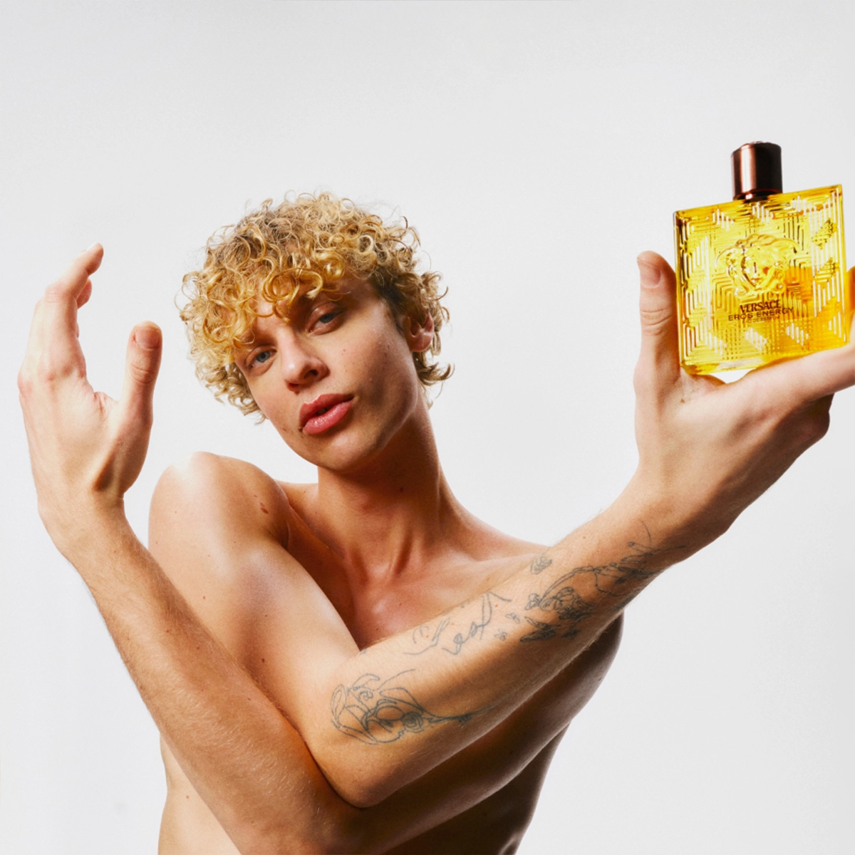 Person med kort krøllete hår holder opp en gul parfymeflaske med teksten "Versace Eros Energy" og Versace-logoen på forsiden. Flasken er transparent med gyllen væske og svart kork mot en lys bakgrunn.