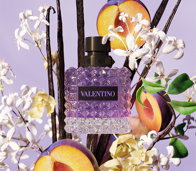 To Valentino-parfymeflasker i lilla toner omgitt av blomster og frukt. Øverst en svart sirkel med teksten «NEW!».