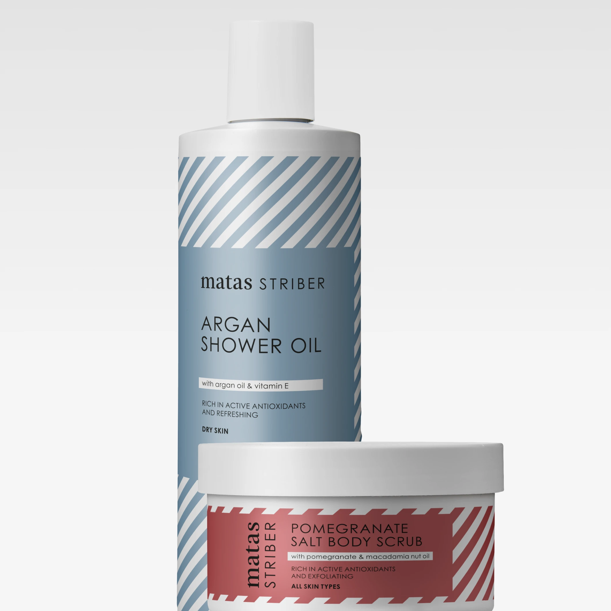 Två kroppsprodukter från matas STRIBER: “ARGAN SHOWER OIL” i en blå flaska och “POMEGRANATE SALT BODY SCRUB” i en burk med röd etikett. På förpackningarna syns texter som “with argan oil & vitamin E”, “RICH IN ACTIVE ANTIOXIDANTS AND REFRESHING”, “DRY SKIN” samt “with pomegranate & macadamia nut oil”.