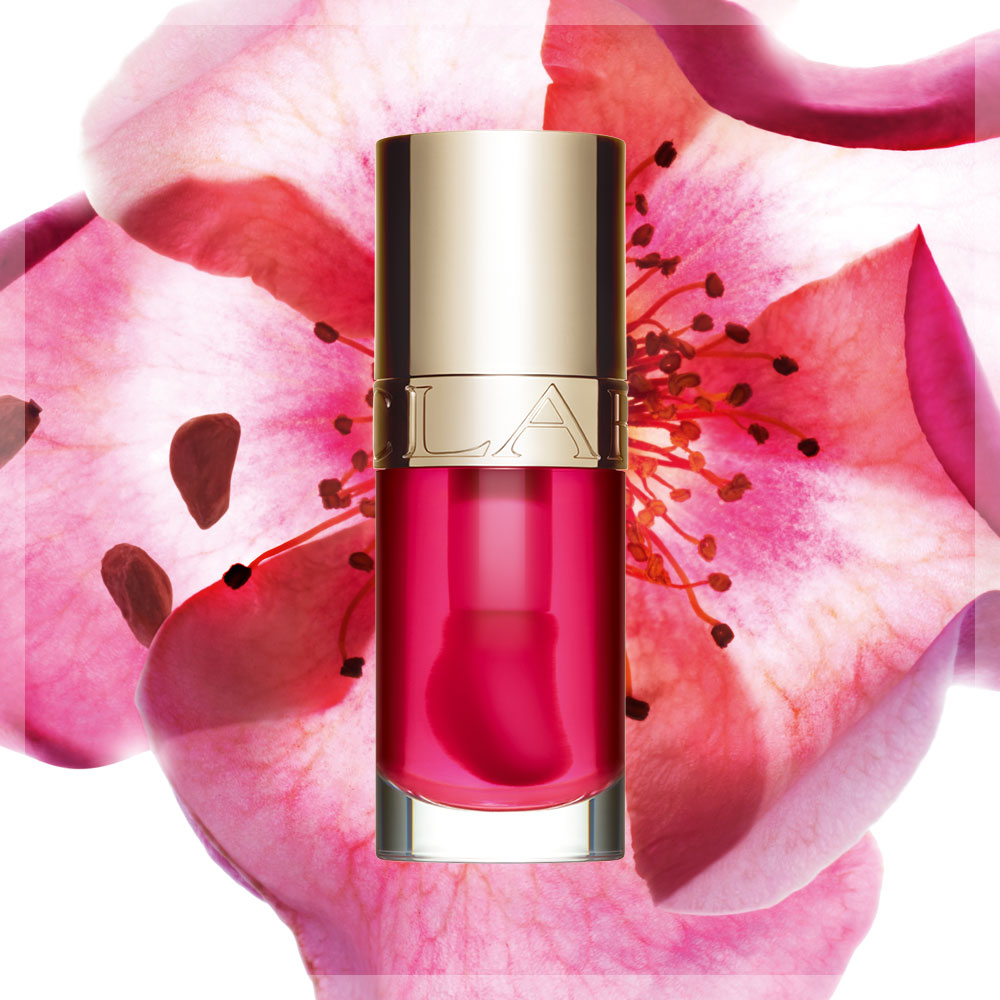 Produktflaska från Clarins med röd vätska, placerad framför en stor rosa blomma och små frön mot en orange bakgrund.