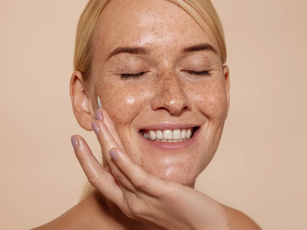 Smilende kvinne med fregner smører hudpleiekrem på kinnet med lukkede øyne.