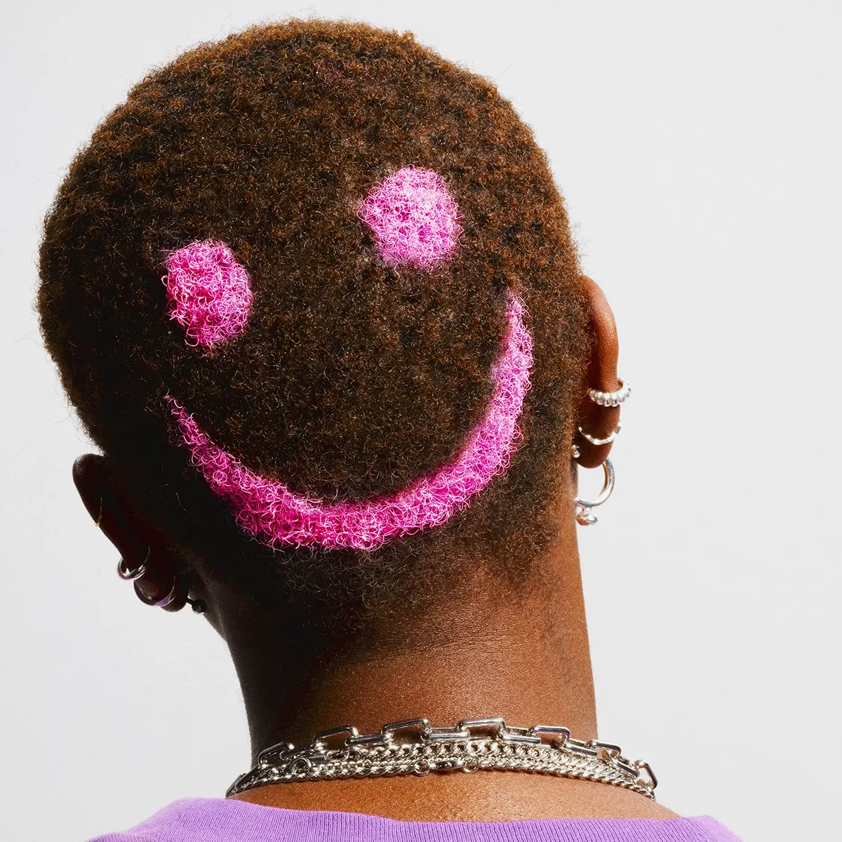 Person bakfra med snauklipt hår og hodet på skrå, mot en hvit bakgrunn. Håret er farget med en rosa smiley i bakhodet.