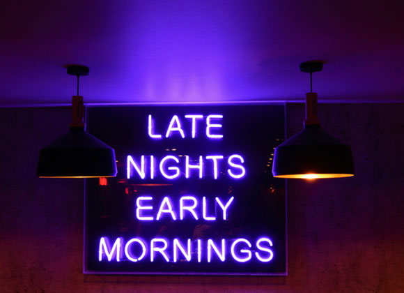 Neonvalokyltti, jossa lukee "Late Nights Early Mornings" pimeässä huoneessa, valaistuna kahdella roikkuvalla lampulla