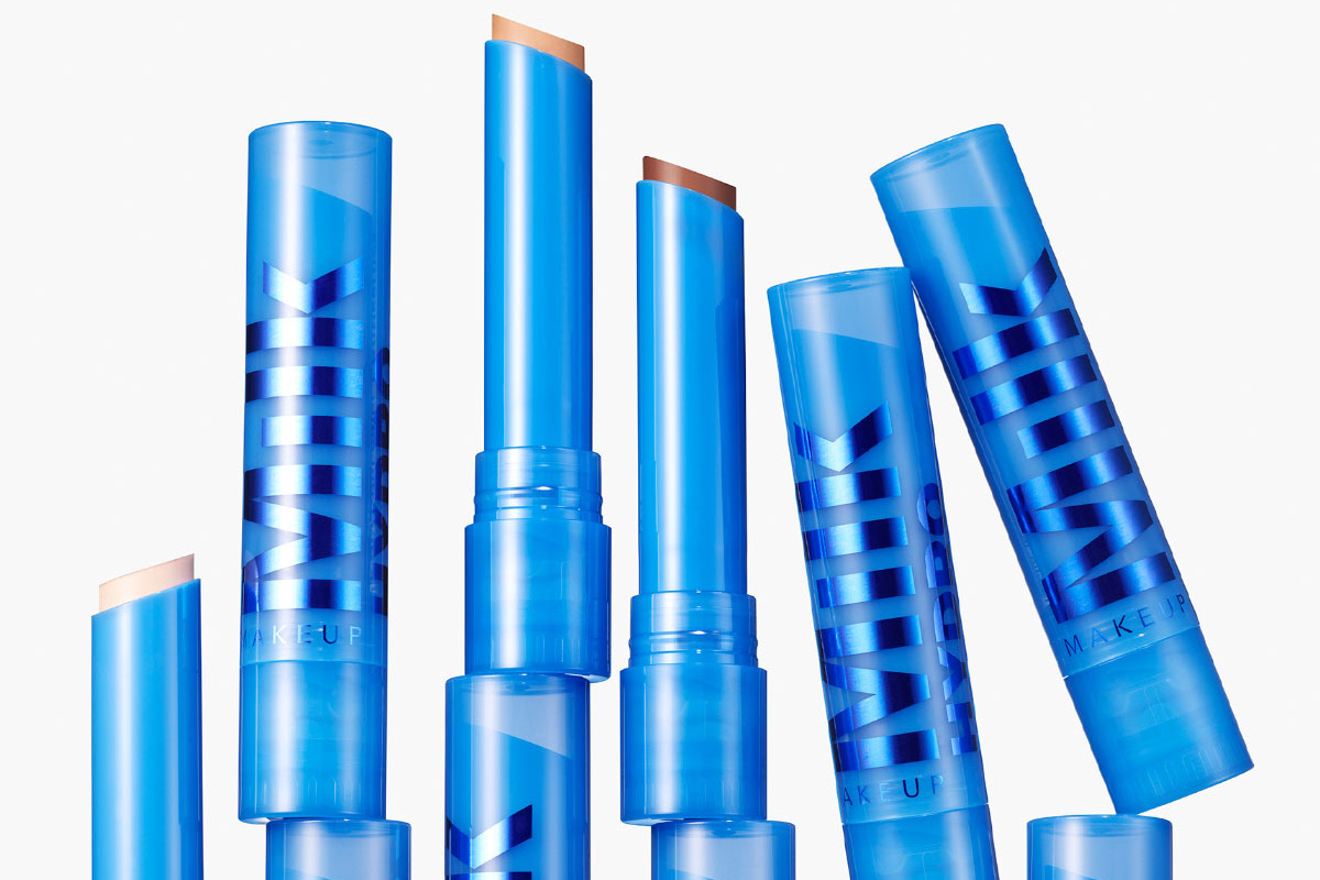 Översikt av flera nyanser av MILK MAKEUP krämiga sminkstift i blå förpackning, arrangerade uppifrån så att färgerna syns från ljusa till mörkare toner. Till höger visas öppnade stift med texten “MILK MAKEUP” och en svart cirkel med texten “NEW!”.