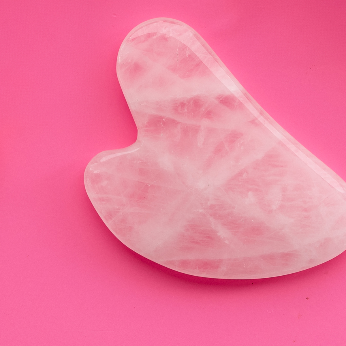 Rosa hjerteformet gua-sha-stein mot cerise bakgrunn.