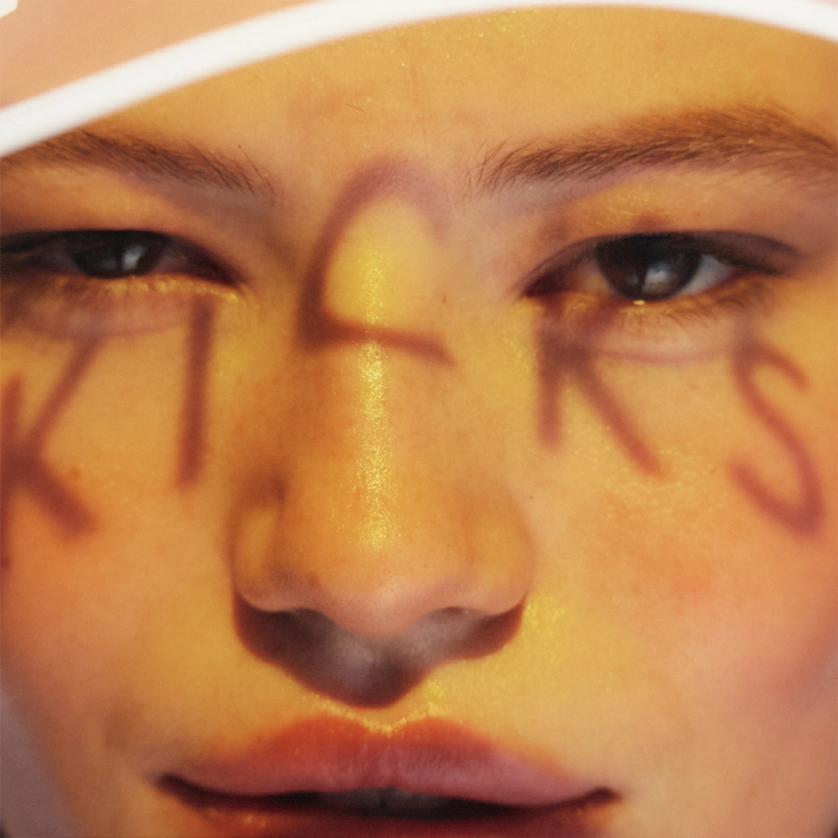 Nærbilde av ansikt med varm, gyllen sminke og glød i huden, med bokstavene "KICKS" malt over ansiktet. Fokus på øyne, hudfinish og kreativ makeup-look.