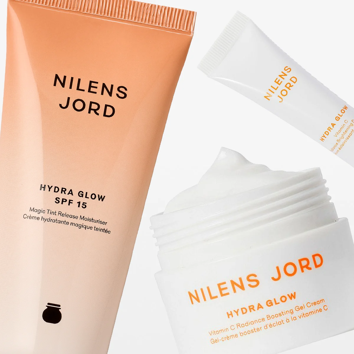 Tre hudpleieprodukter fra Nilens Jord i serien Hydra Glow vises mot en lys bakgrunn. Bildet viser en fuktighetskrem med SPF 15, en gelkrem i krukke og en øyekrem. Produktene har en beige og hvit design og gir en følelse av fukt, glød og naturlig skjønnhet.