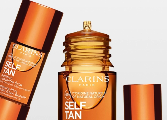 Närbild av bruntonade Clarins self tan-produkter med texten “CLARINS PARIS”, “SELF TAN” och “99% D’ORIGINE NATURELLE / OF NATURAL ORIGIN” på flaskorna mot ljus bakgrund. Flera förpackningar i glänsande kopparfärg visar brun utan sol-produkter för ansikte och kropp.