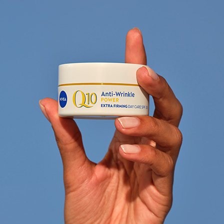En hånd holder en boks med Nivea mot en klarblå himmel. Krukken er hvit med blå og gul tekst som viser produktnavnet. Hånden er elegant plassert, noe som fremhever produktet mot den enkle bakgrunnen.