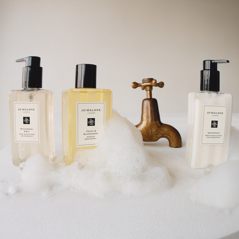 Tre flasker med Jo Malone-produkter plassert på en skummende overflate foran en gammeldags kran.