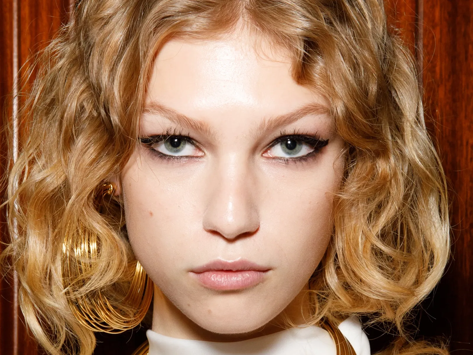 En person med lockigt blont hår, fotograferad framifrån mot en träpanel.