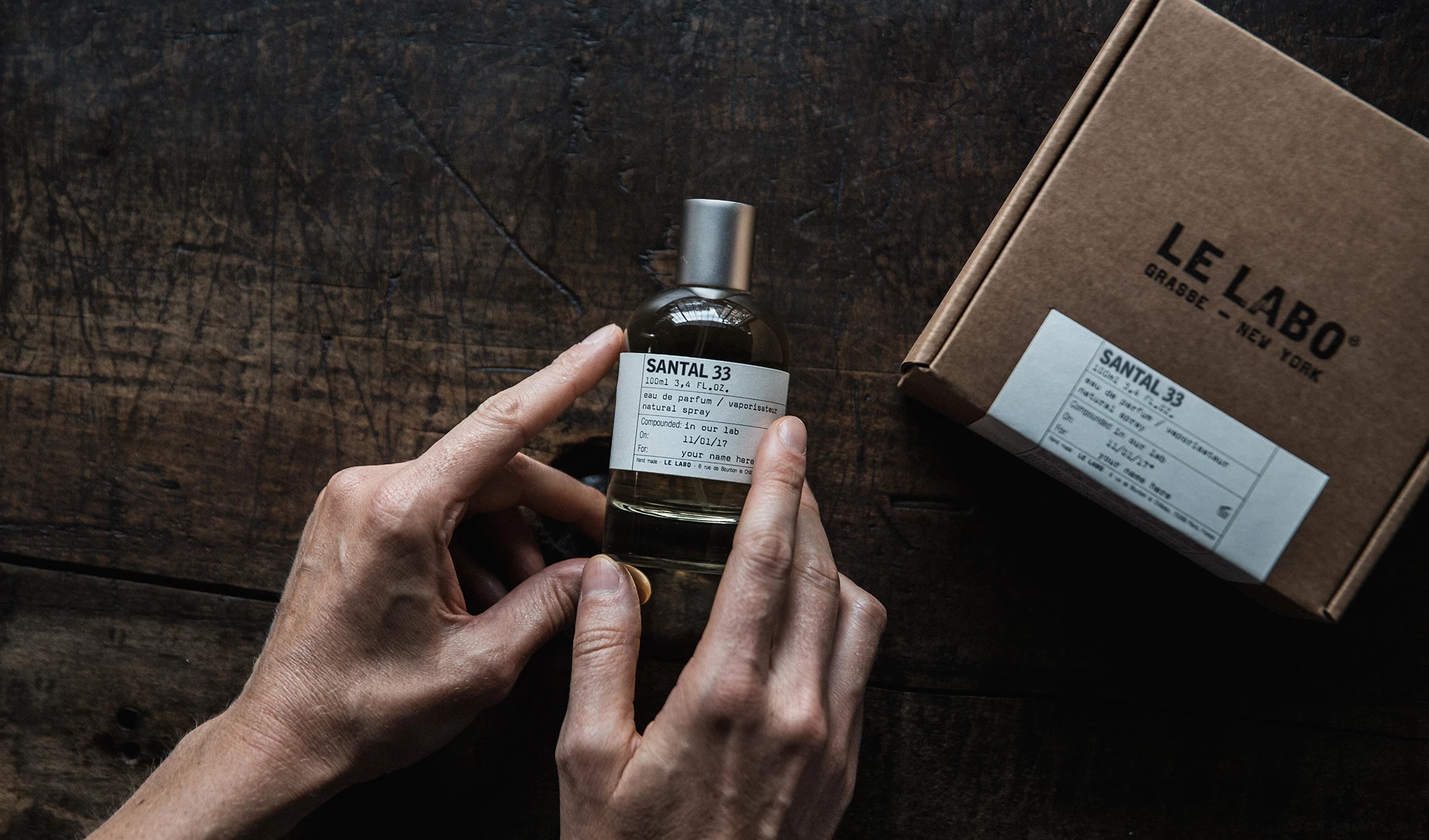 En person holder en flaske med parfymen Santal 33 fra Le Labo. Flasken har en hvit etikett med tekst og står på et mørkt trebord. Til høyre for flasken står en brun pappeske med Le Labos logo og en etikett som matcher etiketten på flasken.