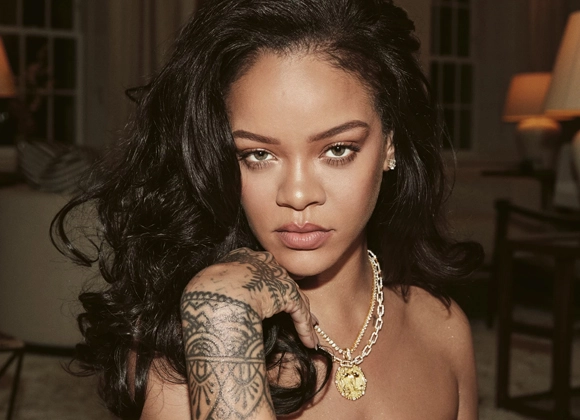 Rihanna katsoo kameraan kaulakoru päässään ja näyttää tatuoinneilla varustettua kättään. Istuminen olohuoneessa.