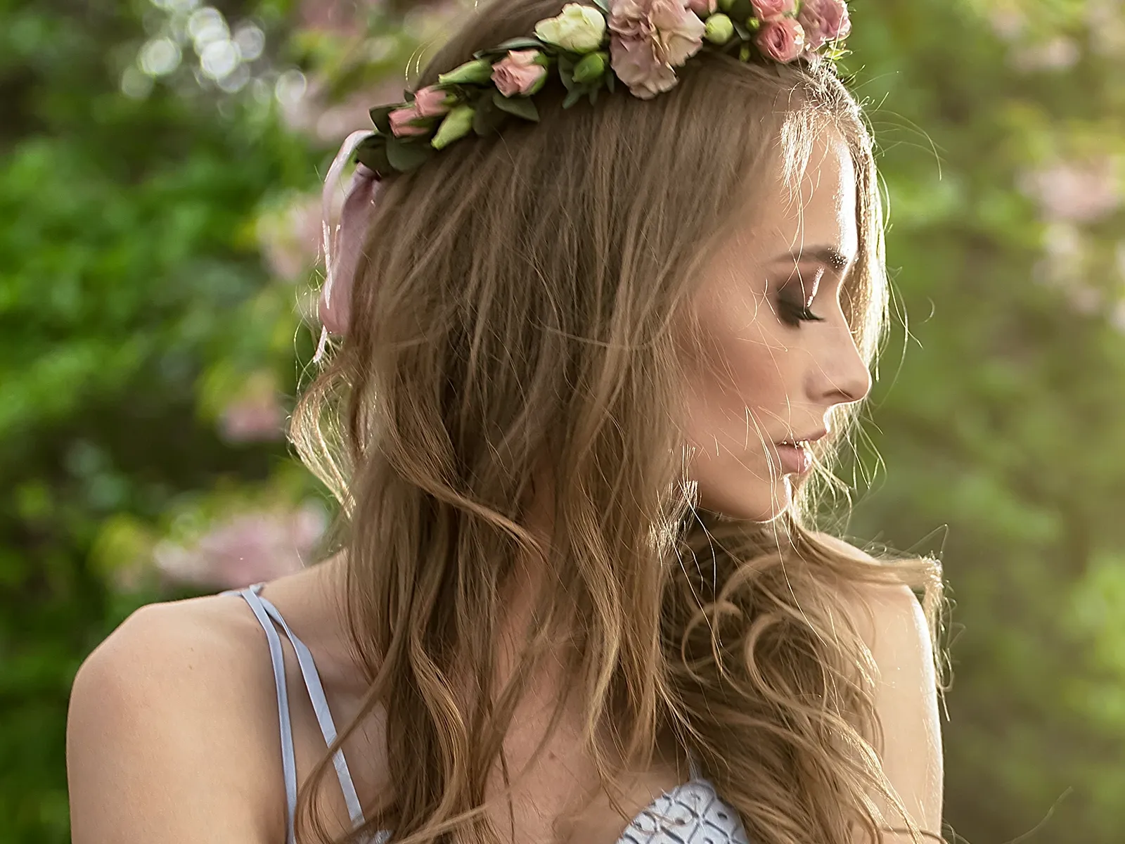 En kvinna med långt hår och en blomkrans på huvudet, i profil framför gröna träd.