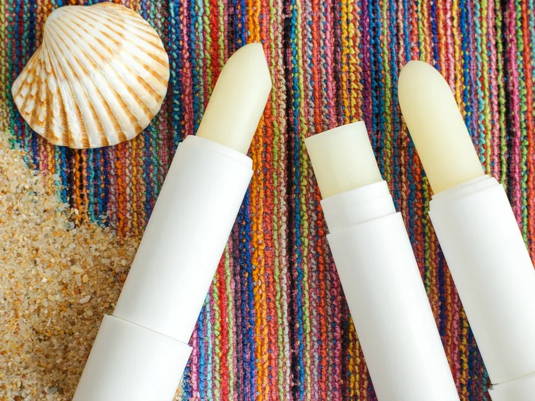 Leppepomadestifter på et fargerikt vevd stoff med skjell og sand.