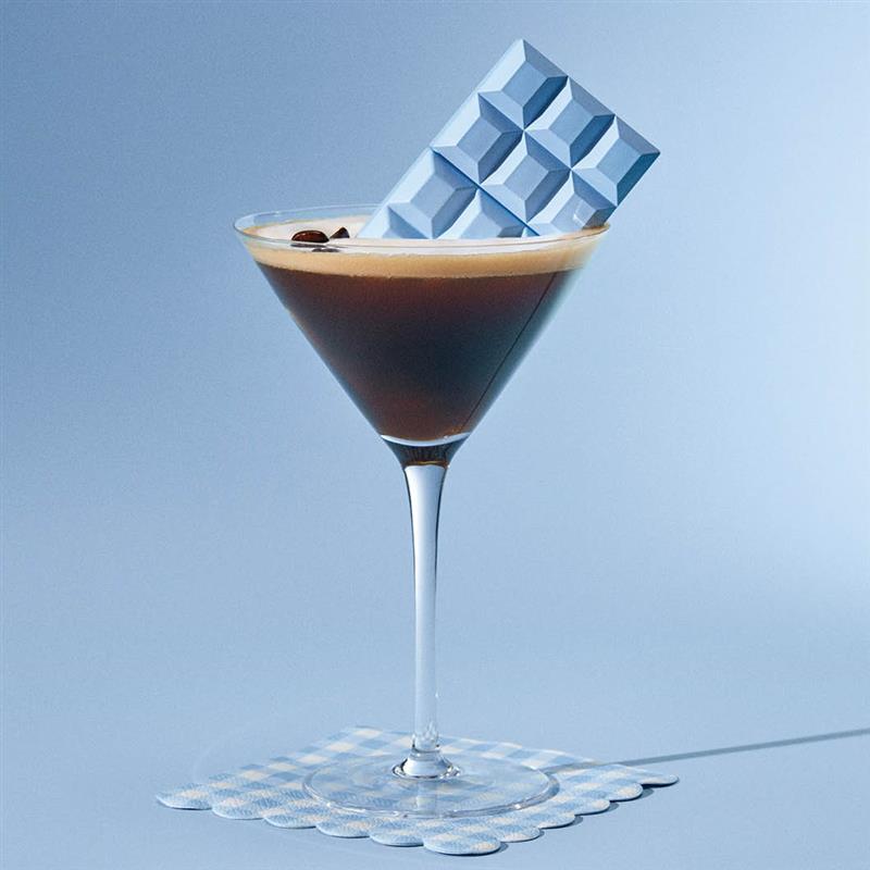 Et martini-glass med en mørk drikke og en lyseblå sjokoladebit. Under glasset er det en lyseblå rutete bordbrikke.