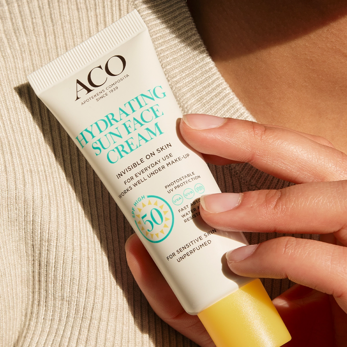 Närbild av en hand som håller en tub ACO Hydrating Sun Face Cream SPF 50 med texten “Invisible on skin”, “For everyday use works well under make-up”, “High protection 50” och “For sensitive skin unperfumed”, mot en neutral bakgrund. Fokus på solskydd för ansiktet och hudvård.