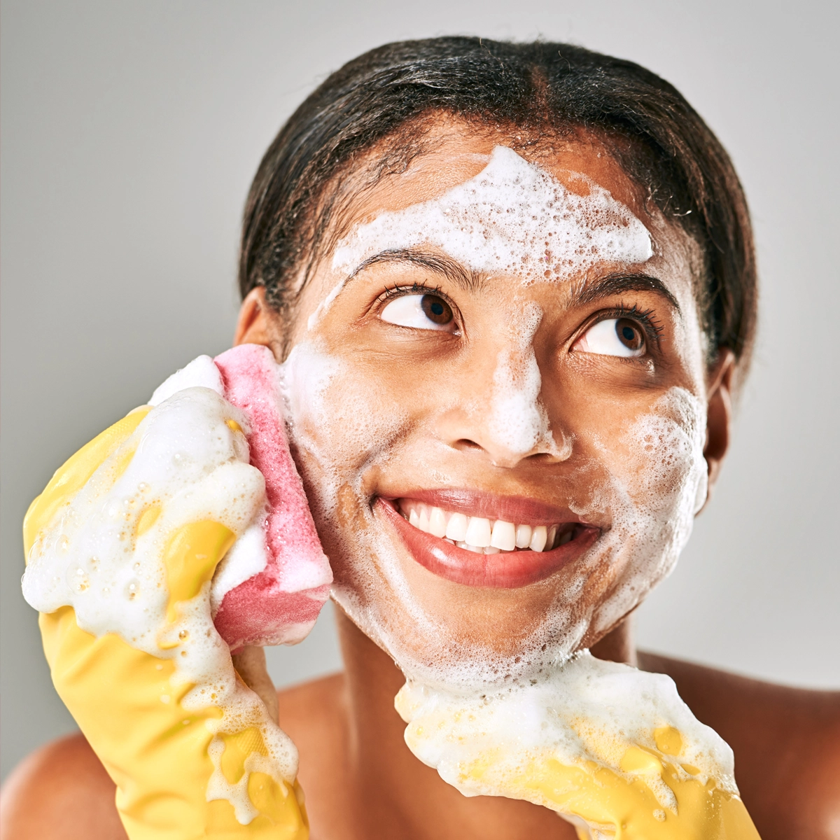 Person med uppsatt hår tvättar ansiktet med rikligt lödder. Hen håller en rosa och gul tvättsvamp mot kinden och bär gula rengöringshandskar. Vitt skum täcker panna, kinder och haka mot ljus bakgrund.