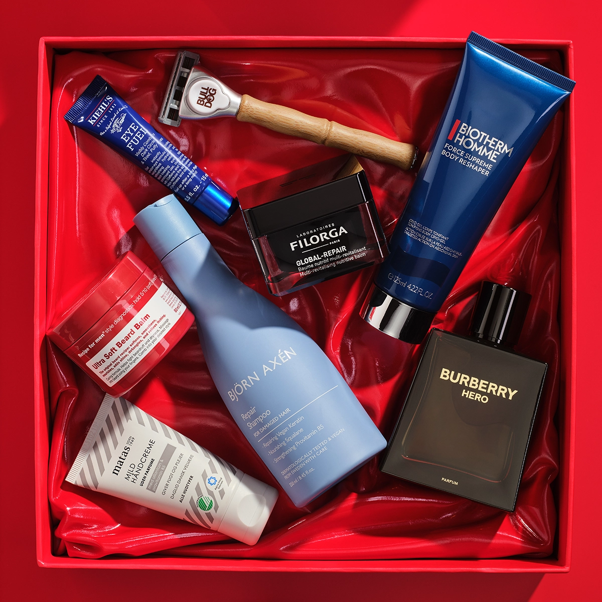 Bilde som viser flere skjønnhetsprodukter arrangert i en rød gaveeske. Produktene inkluderer en blå tube med Kiehl's Eye Fuel, en barberhøvel fra Bulldog, en svart boks med Filorga Global-Repair, en blå tube med Biotherm Homme Force Supreme Body Reshaper, en svart flaske med Burberry Hero parfyme, en blå flaske med Björn Axén Repair Shampoo, en hvit tube med Matas Men Ansiktskrem, og en rød boks med Recipe for Men Ultra Soft Beard Balm. Bakgrunnen er rød.