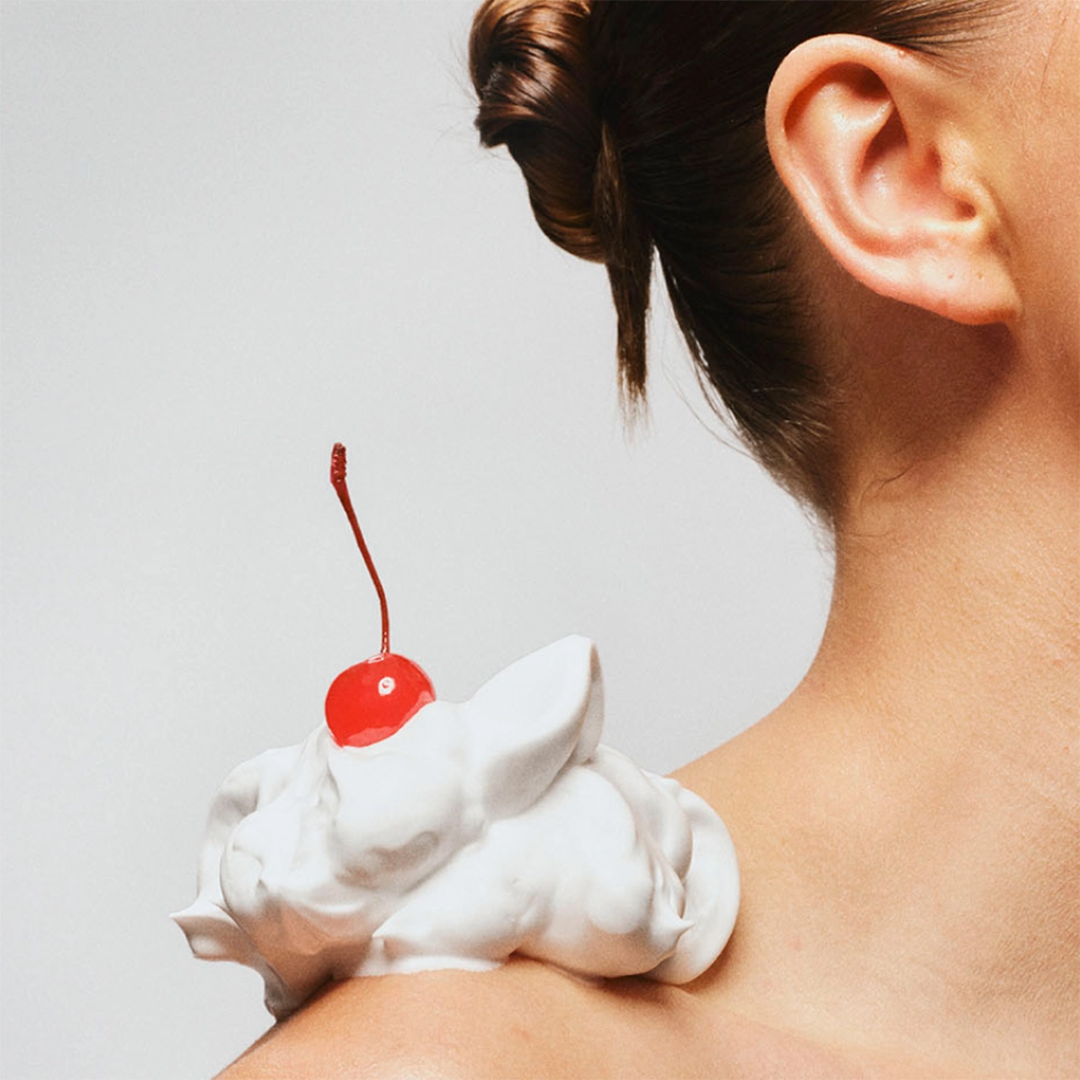 Lähikuva paljaasta olkapäästä, jonka päällä on nokare valkoista voidetta ja sen päällä punainen kirsikka, neutraalia taustaa vasten.