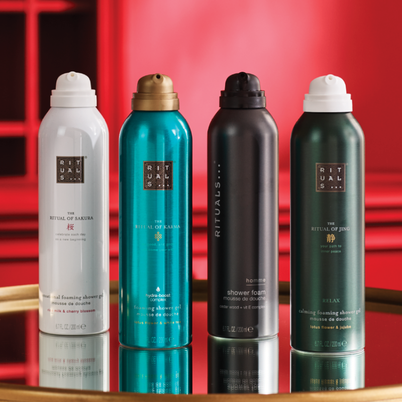 Fyra Rituals duschgelflaskor placerade bredvid varandra framför en röd bakgrund. Flaskorna har färgerna vit, turkos, svart och mörkgrön.