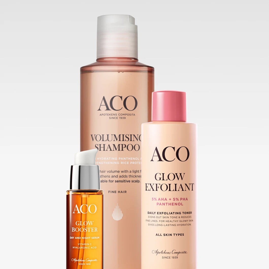 Bild som visar tre ACO produkter: en brun flaska med ACO Volumising Shampoo, en rosa flaska med ACO Glow Exfoliant, och en orange flaska med ACO Glow Booster Day and Night Serum. Bakgrunden är vit.