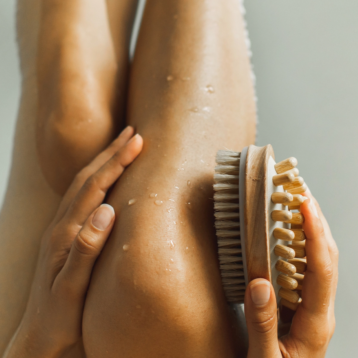 En person använder en torrborste för kroppsvård på ett ben med vattendroppar på huden. Träborsten med naturfärgade borst hålls mot benet i en minimalistisk spa- och wellnessmiljö med fokus på exfoliering och body care.