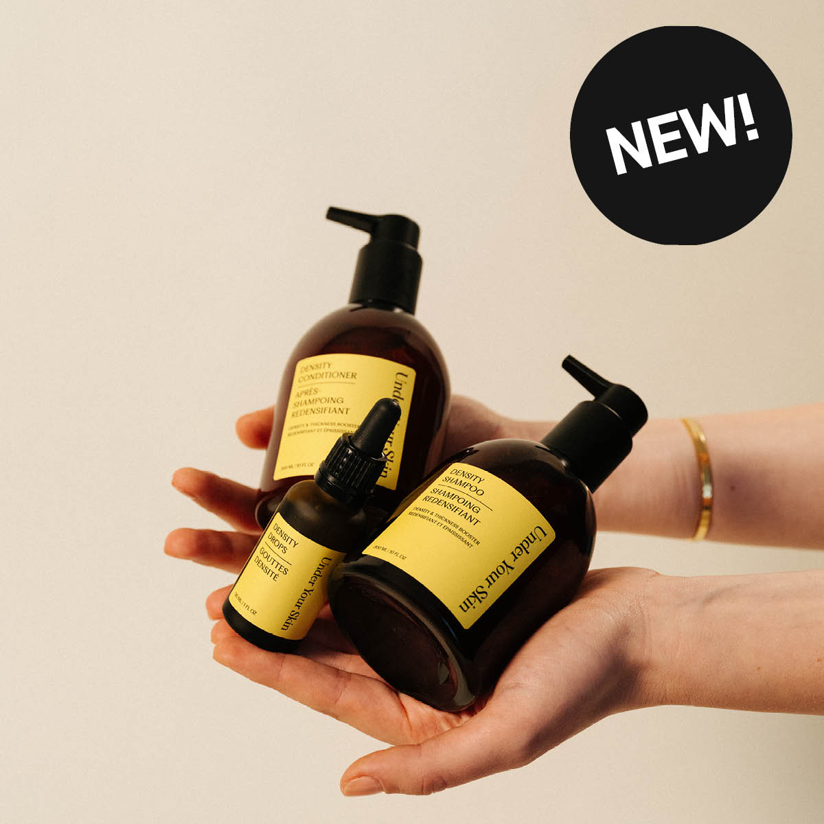 Tre hårpleieprodukter fra Under Your Skin holdes i hendene mot beige bakgrunn: “Density Shampoo”, “Density Conditioner” og “Density Drops”. Gullfargede etiketter og svart sirkel med teksten “NEW!” vises i bildet.