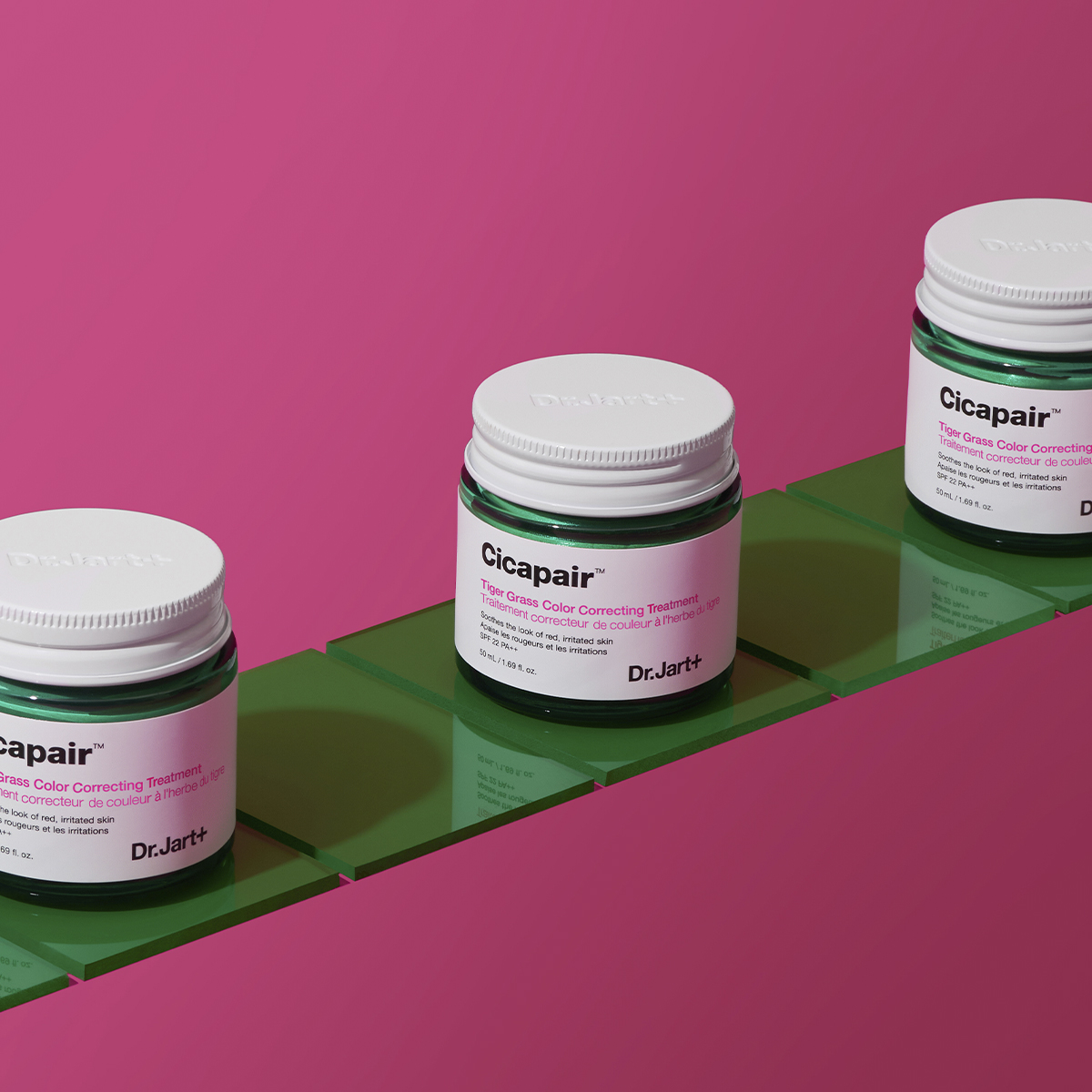 Tre krukker med Dr.Jart+ Cicapair Tiger Grass Colour Correcting Treatment, plassert på grønne fliser mot en rosa bakgrunn.