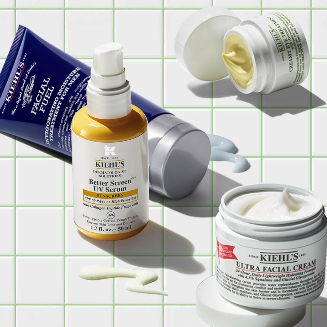 Flere hudpleieprodukter fra Kiehl’s ligger på en hvit flisbakgrunn med grønne linjer, inkludert “Better Screen UV Serum SPF 50 PA++++”, “Ultra Facial Cream” og “Facial Fuel”. Kremer og lotion er smurt ut ved siden av produktene.