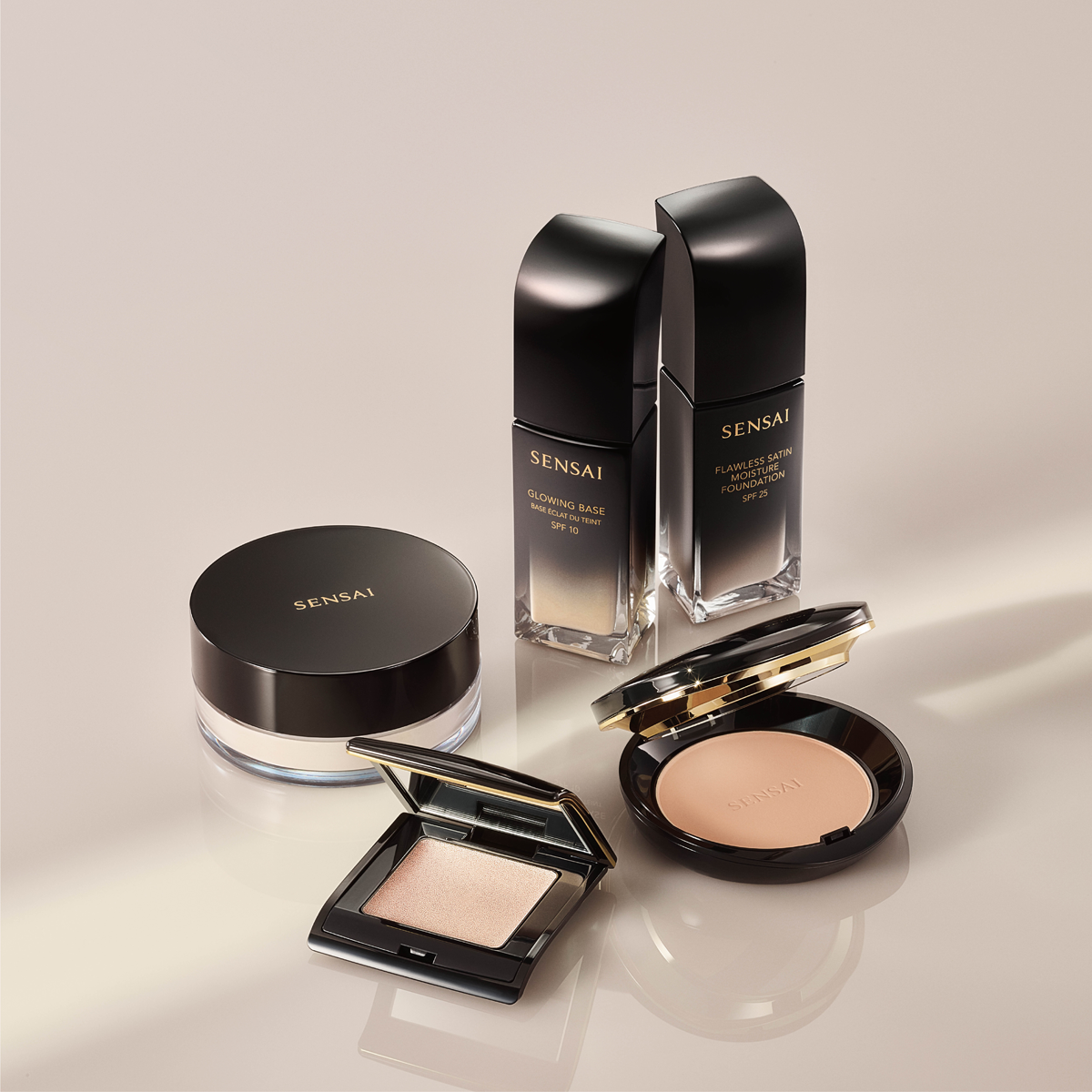 Useita SENSAI-meikkituotteita esillä beigen sävyisellä taustalla, mukana Glowing Base SPF 10, Flawless Satin Moisture Foundation SPF 25, puuteri, highlighter ja compact foundation. Tuotteissa on musta ja kullanvärinen design peilaavilla yksityiskohdilla, jotka luovat ylellisen vaikutelman premium foundation- ja glow-finish makeup -tuotteista.