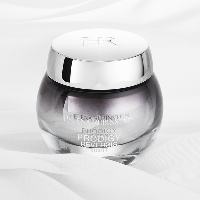 En elegant krukke med Helena Rubinstein Prodigy Reversis Night Cream mot en hvit bakgrunn med myke stofffolder. Krukken er rund og gjennomsiktig med en sølvfarget kork med inngravert «HR». Fargen på kremen er en subtil lilla nyanse, og teksten på krukken viser produktets navn og varemerke. Designet er luksuriøst og sofistikert.