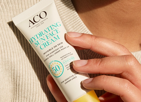 Närbild av en hand som håller en tub ACO Hydrating Sun Face Cream SPF 50 med texten “Invisible on skin”, “For everyday use works well under make-up”, “High protection 50” och “For sensitive skin unperfumed”, mot en neutral bakgrund. Fokus på solskydd för ansiktet och hudvård.