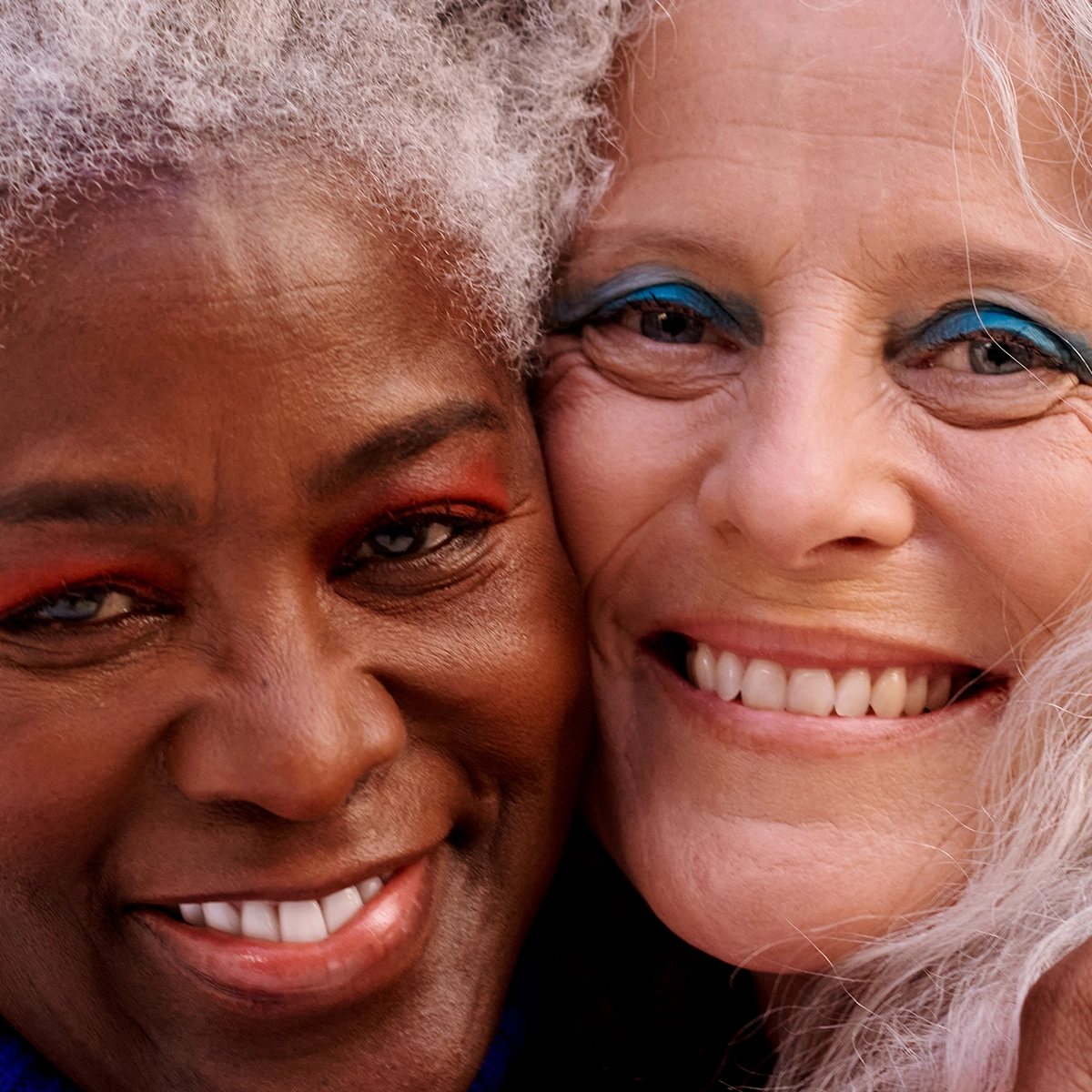 Lähikuva kahdesta iäkkäästä henkilöstä, jotka hymyilevät kameraan posket lähekkäin. Molemmilla on vaalea silmämeikki sinisen ja oranssin sävyissä sekä harmaat hiukset. Kuva on rajattu niin, että vain kasvot ja osa hiuksista näkyvät.